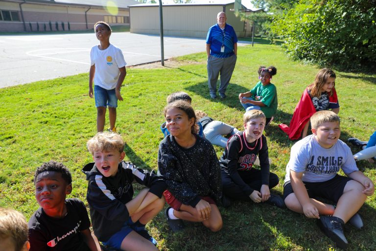 Group of children sitting on grass in a schoolyard, with an adult standing nearby.