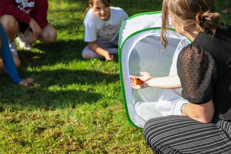 Woman in black shirt kneeling on grass holding net with insects inside, surrounded by children observing.