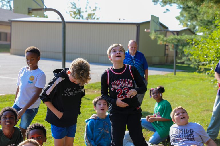 Children gathered in a park, posing for a picture, with adults supervising them.