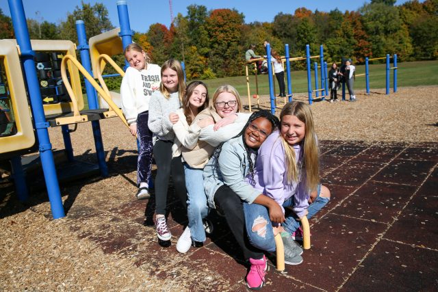 Six girls on playground equipment playing with fall trees in the background
