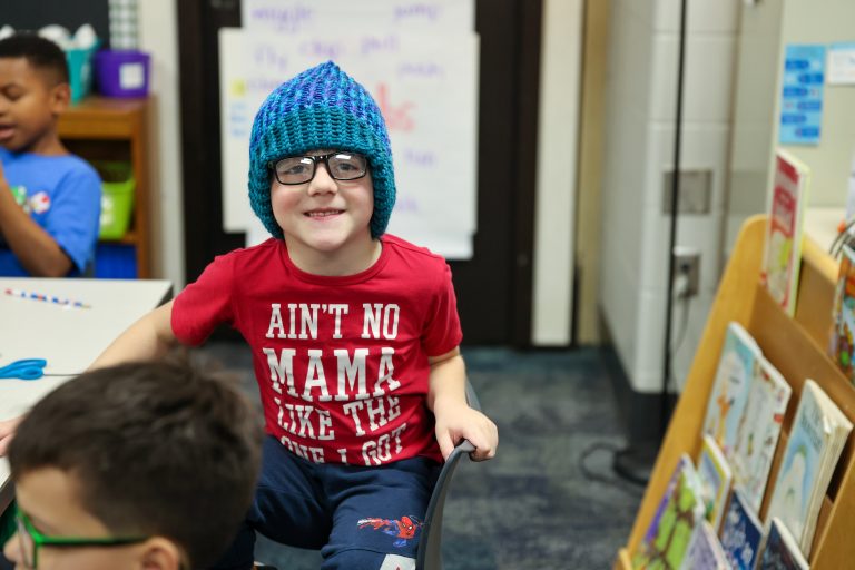 Young boy in a classroom setting wearing glasses and a blue hat, sitting at a desk.