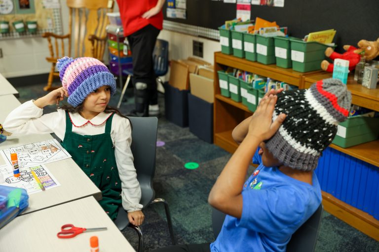 Two children wearing knitted hats, sitting at a table in a classroom setting, engaging in an activity.