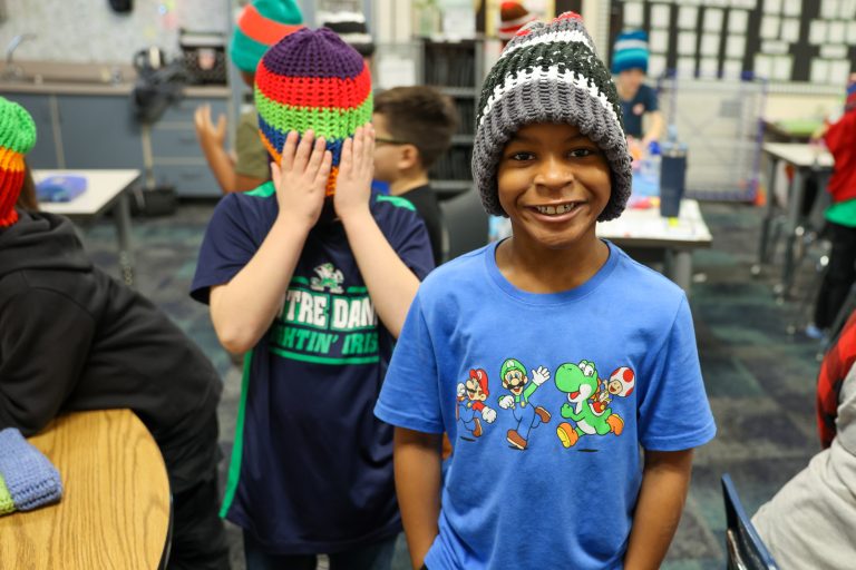 Young boy in blue shirt and knit hat smiling at camera, standing in front of table with other children wearing hats, posing