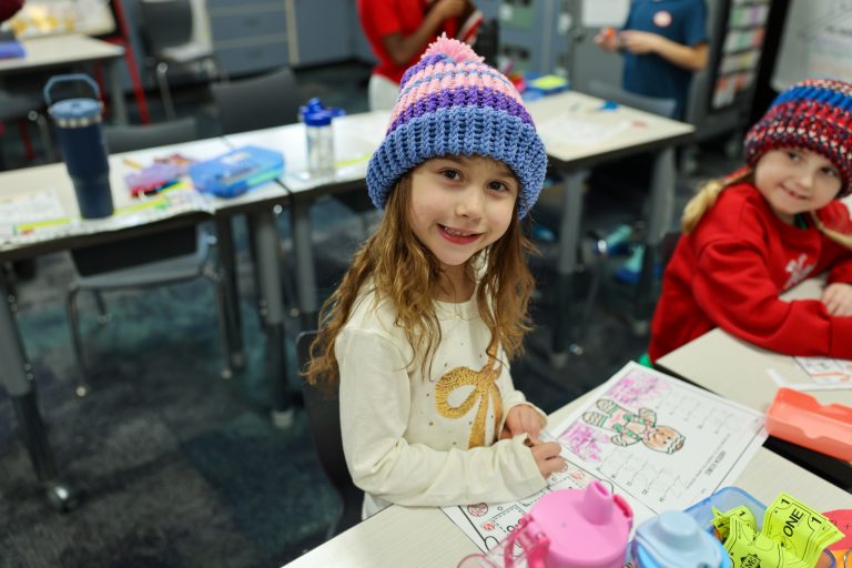 Girl sitting at desk in classroom, smiling at camera, wearing blue hat with pompom and pink stripes.