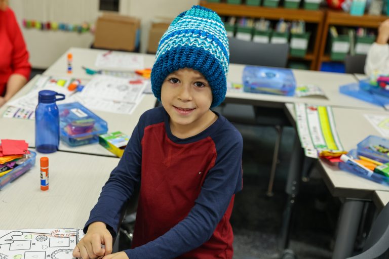Boy wearing blue knit hat at school desk.