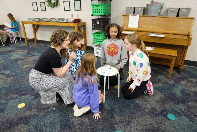 Five people gathered around a small table in a room with blue carpeting, discussing something while one person is seated on
