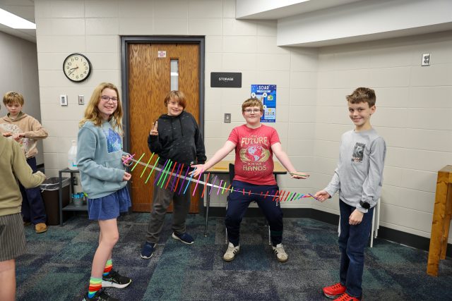 Group of young people in a classroom, posing for a picture while holding a colorful ribbon.