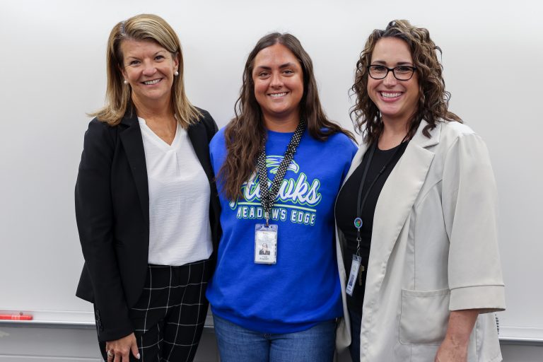 Three women standing in front of a whiteboard, posing for a picture. The woman on the left is wearing a blue sweatshirt with