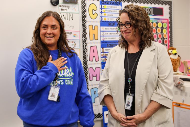 Two women standing in front of a classroom bulletin board. One woman is wearing a blue shirt and has her hands on her chest,