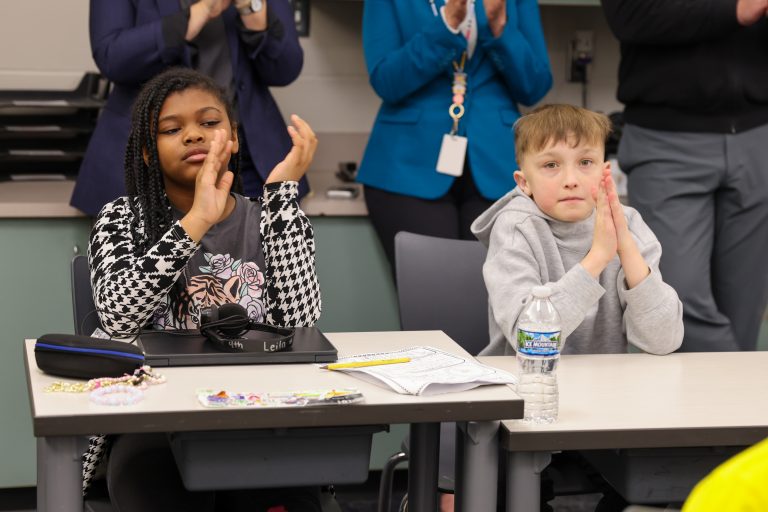 Two children clapping hands at a table in front of an audience, one child is seated and the other stands behind them.