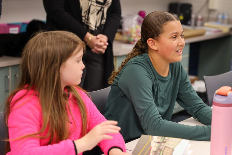 Two young girls sitting at a table in a classroom setting, engaged in an activity. The girl on the left appears to be