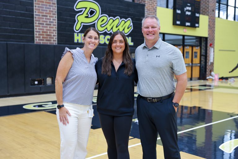 Two women posing in front of a basketball court. One woman is wearing a black shirt and the other is wearing a white shirt.