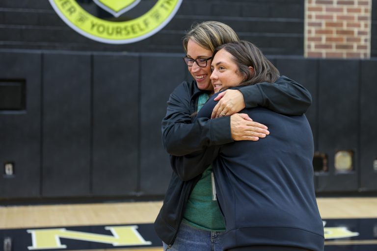 Two people embracing in a gymnasium, one person wearing glasses, both smiling.
