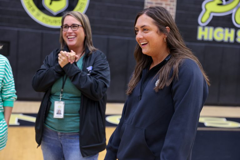 Three people celebrating, two women clapping, one woman smiling broadly, all in a gymnasium with a basketball hoop and team